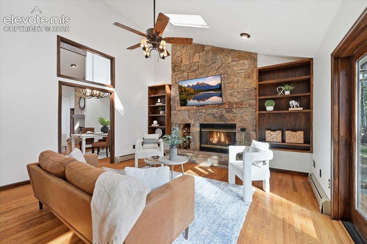 Vaulted ceiling with a skylight and ceiling fan, stone-faced fireplace, hardwood flooring, built-in shelving, and a large doorway with transom window