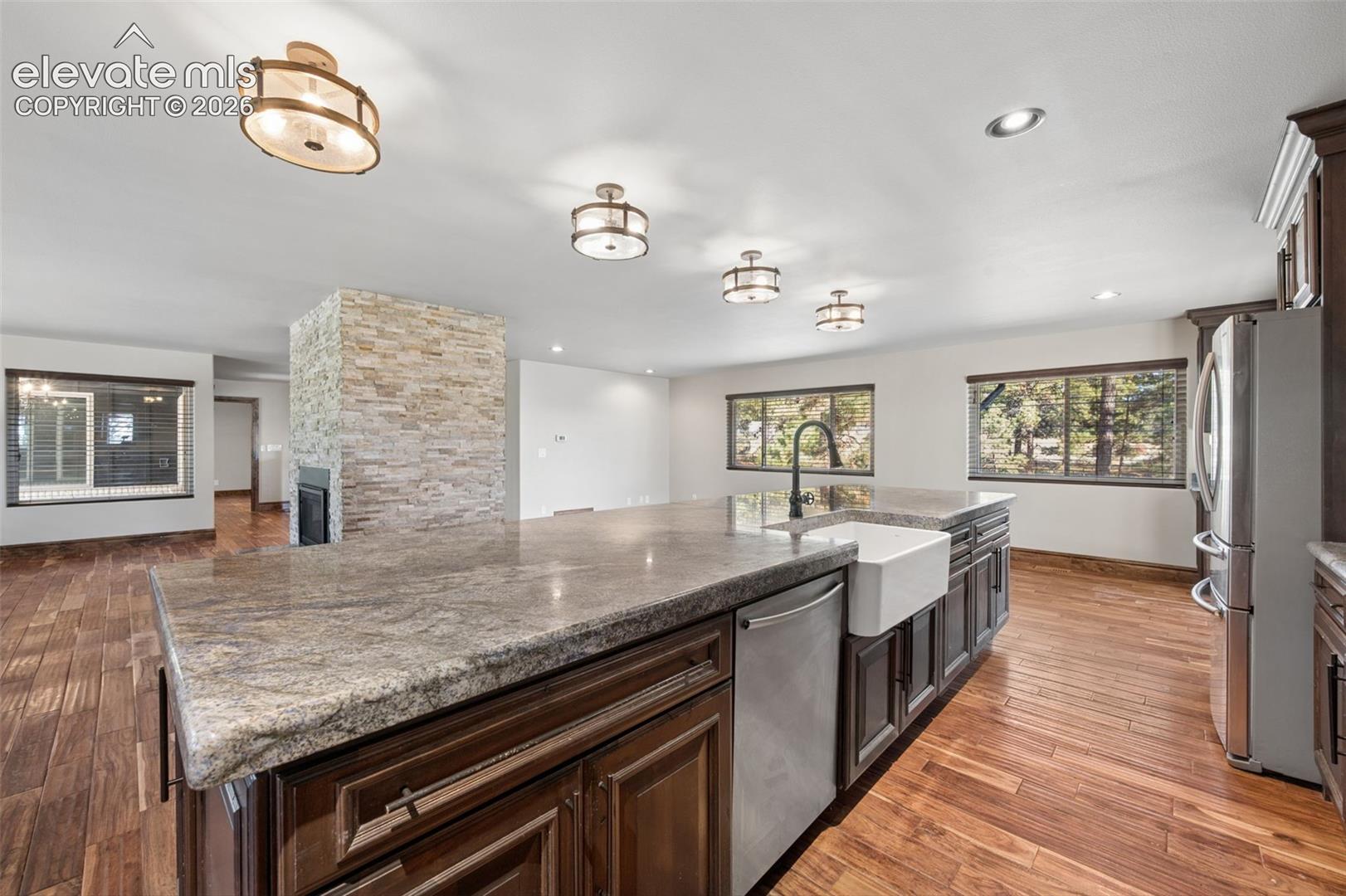 Kitchen featuring a large island with an expansive countertop, farmhouse-style apron sink, matte black faucet, dark cabinetry, and wood-finish flooring, complemented by a floor-to-ceiling stone-faced fireplace