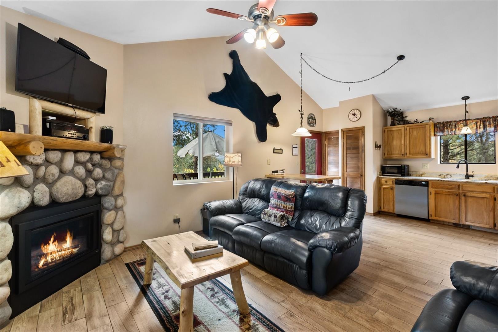 Stone-clad fireplace with wood mantle, wood-finish flooring, vaulted ceiling with ceiling fan, and built-in kitchen cabinetry with countertop