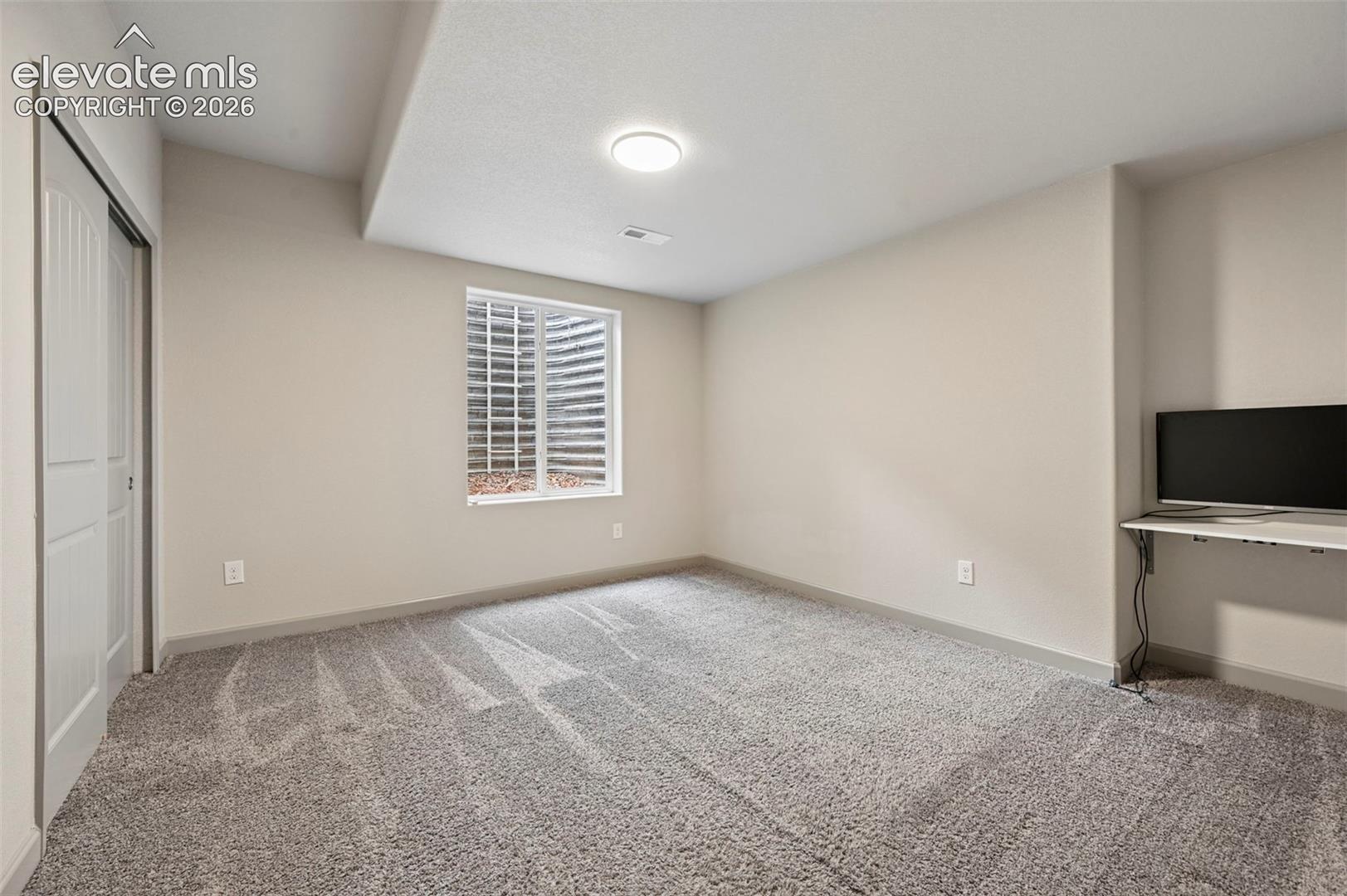 Neutral-toned room featuring carpeted flooring, a recessed window with an egress well, a built-in wall-mounted desk, and a sliding-door closet