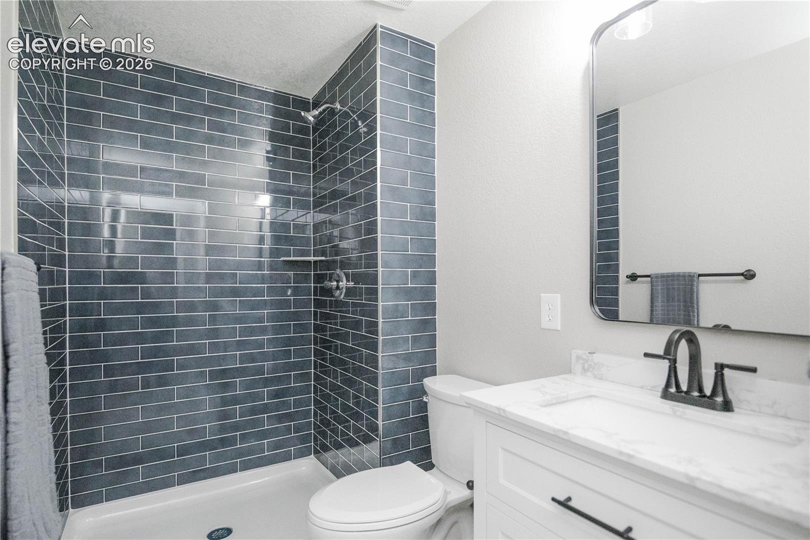 Bathroom featuring a walk-in shower with blue subway tile surround, white vanity cabinet with a stone-top integrated sink, matte black hardware, and an arched wall-mounted mirror