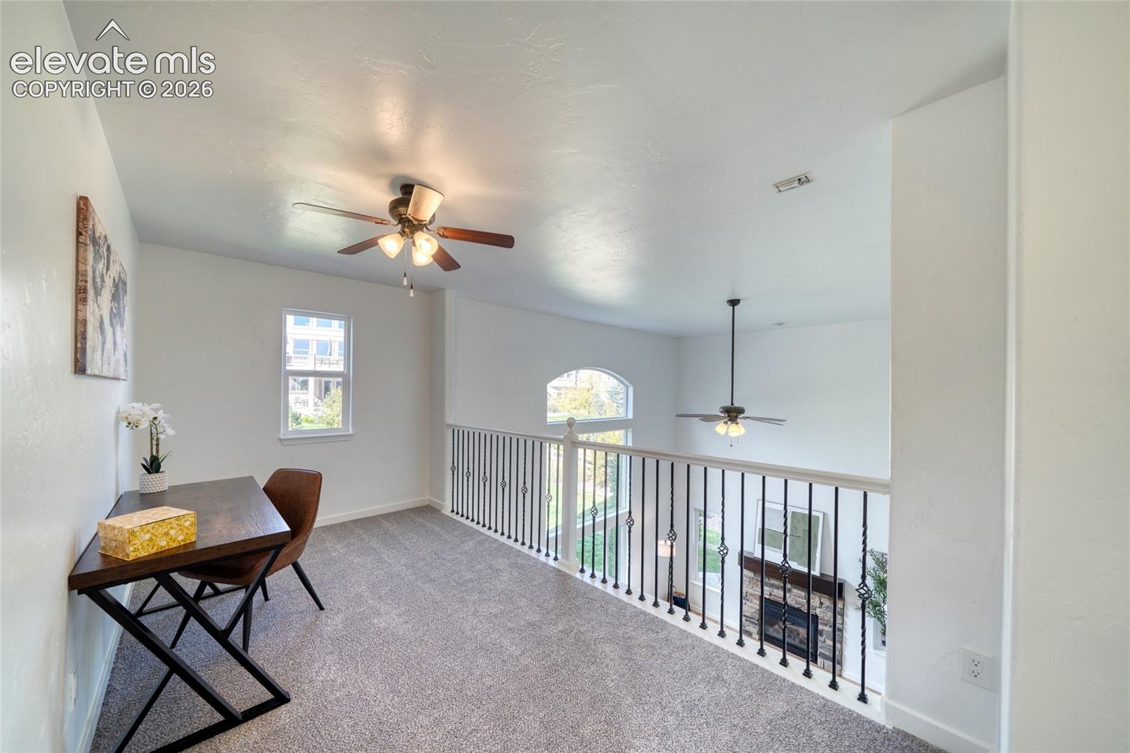 Carpeted open loft area featuring a ceiling fan, a window, and a wrought iron railing overlooking the lower level