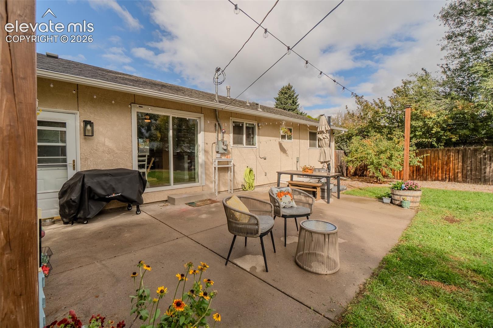Rear exterior featuring a concrete patio, exterior wall-mounted string lighting, sliding glass door, and a wooden privacy fence