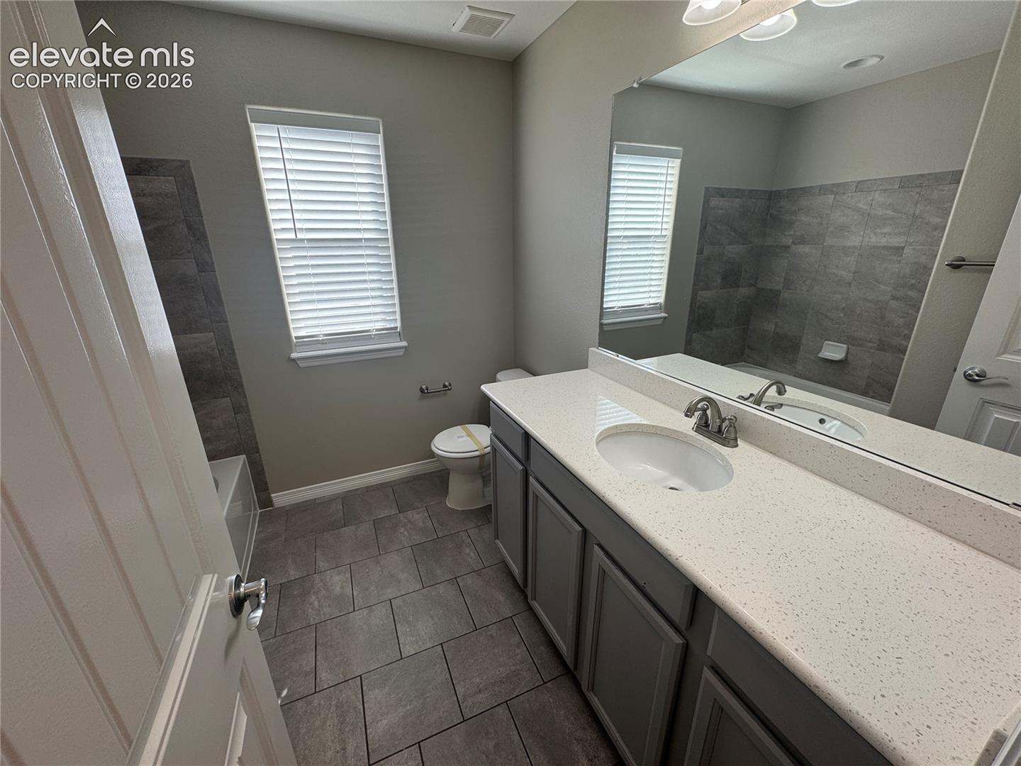 Bathroom featuring a vanity with shaker-style cabinetry and a composite countertop, an oversized wall-mounted mirror, ceramic tile flooring, and a shower-tub combination with stone-finish wall tile