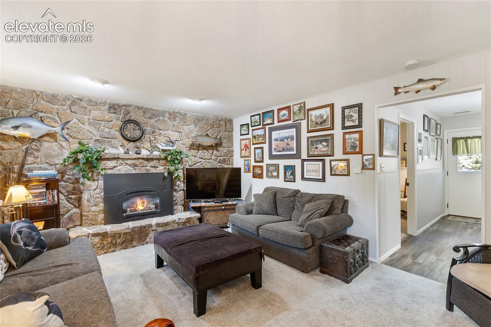 Stone-clad fireplace with a hearth, carpeted flooring, white paneled wall accents, and wood-finish flooring in the hallway
