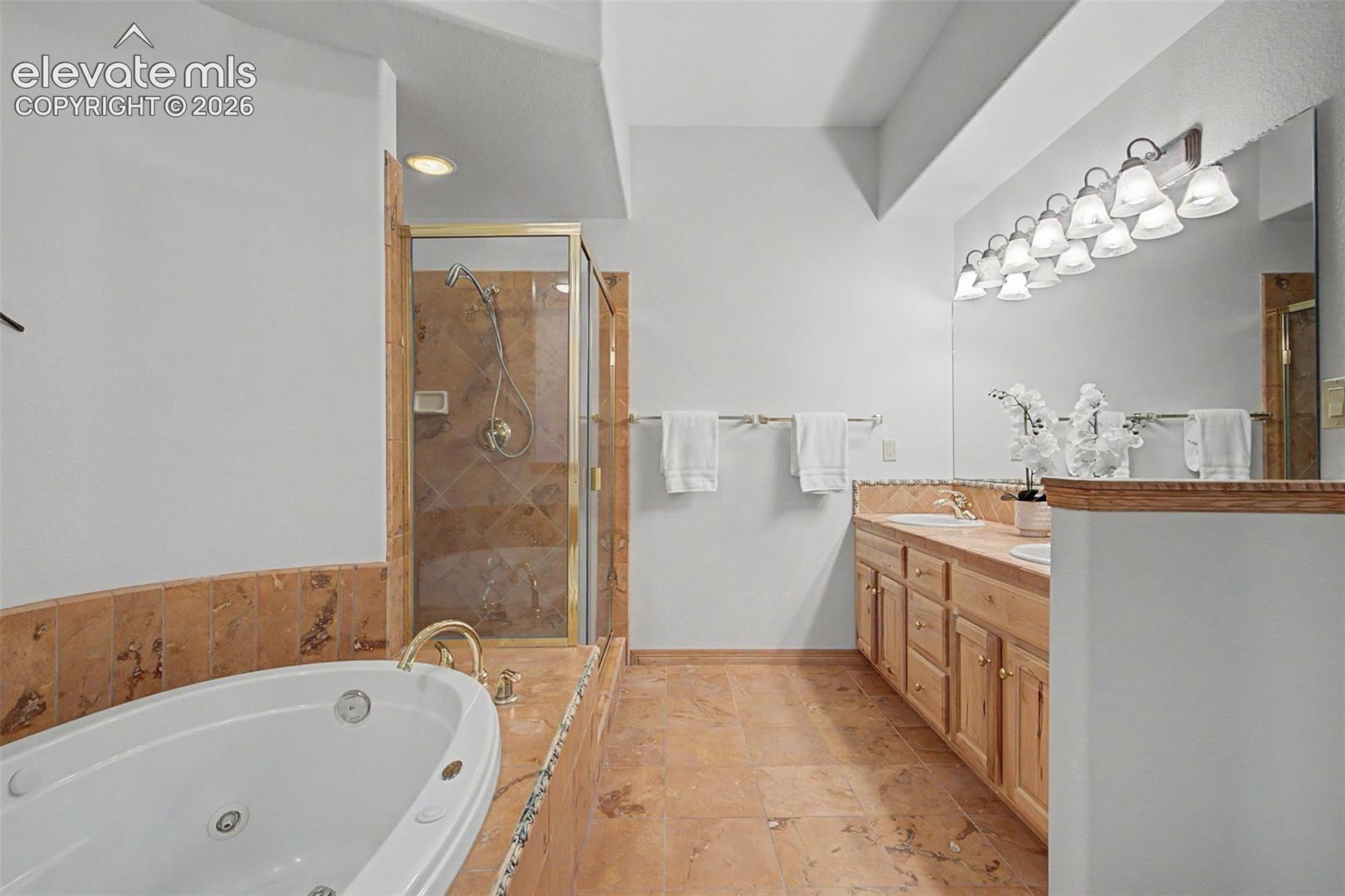 Spacious bathroom featuring a jetted soaking tub, a stand-up shower with glass enclosure, a dual-sink vanity with light wood cabinetry, and extensive tile work on floors and tub surround