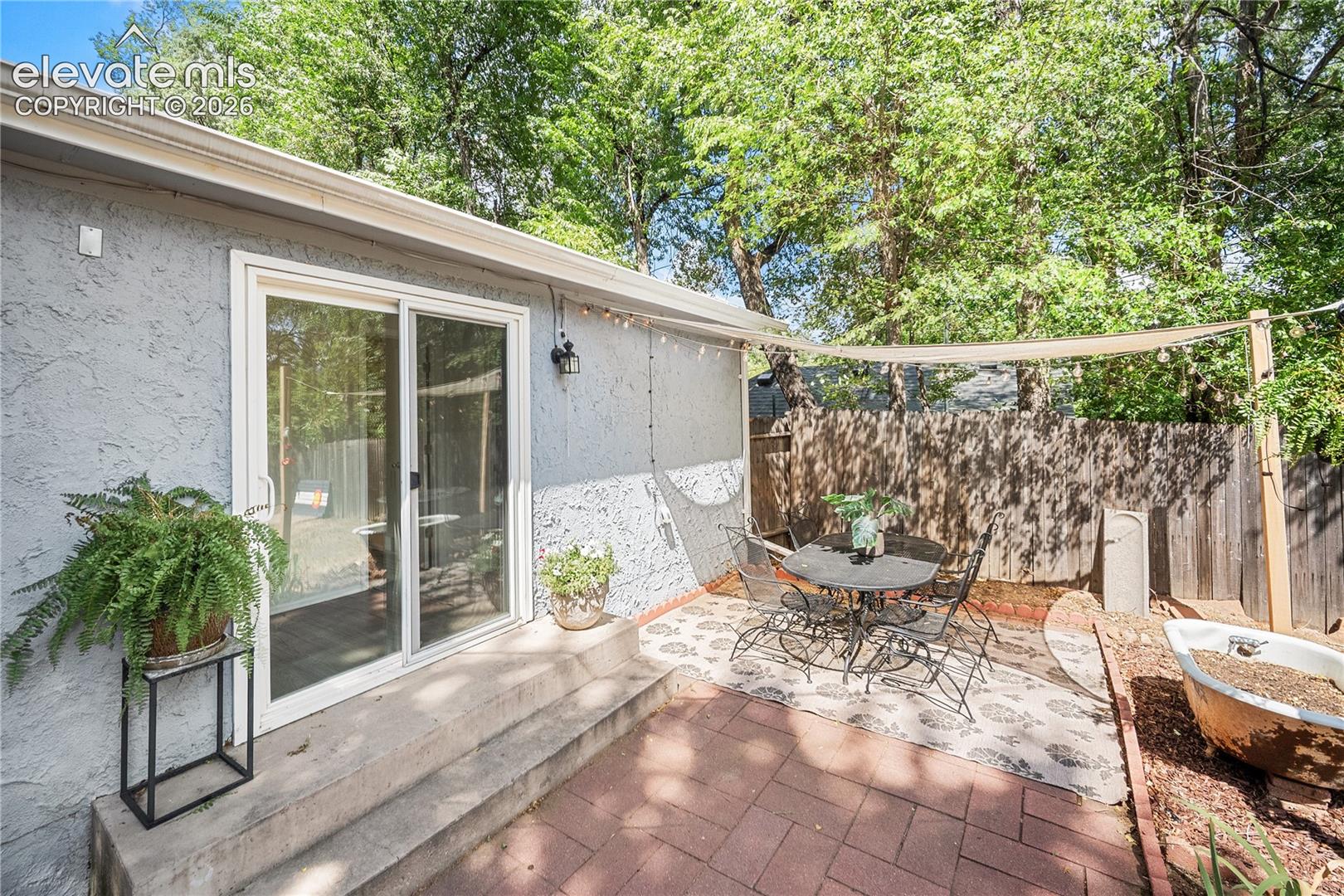 Brick paver patio with a shade sail, bordered by a wooden privacy fence and mature trees