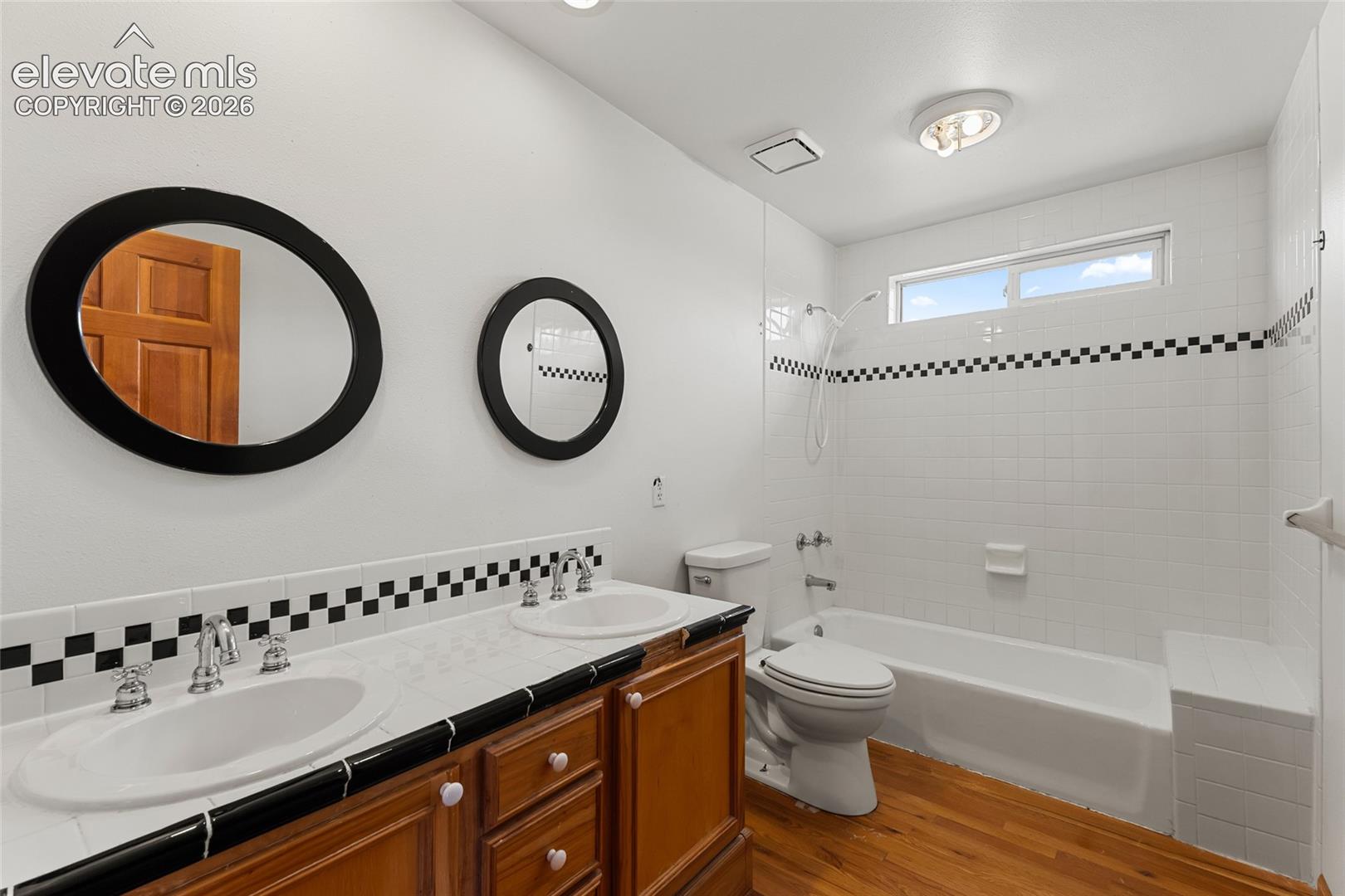 Dual vanity with integrated sinks, wood-finish cabinetry, a built-in bathtub with shower, and tile flooring
