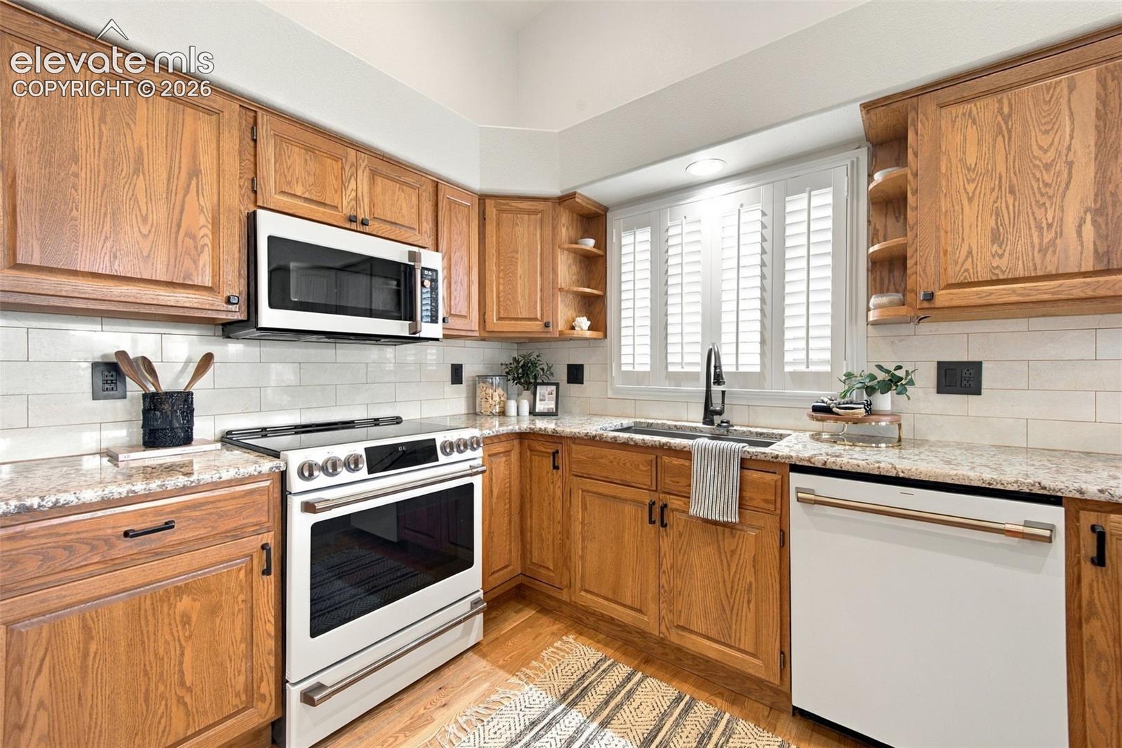 Kitchen featuring wood cabinetry, stone-finish countertops, subway tile backsplash, white appliances, and plantation shutters