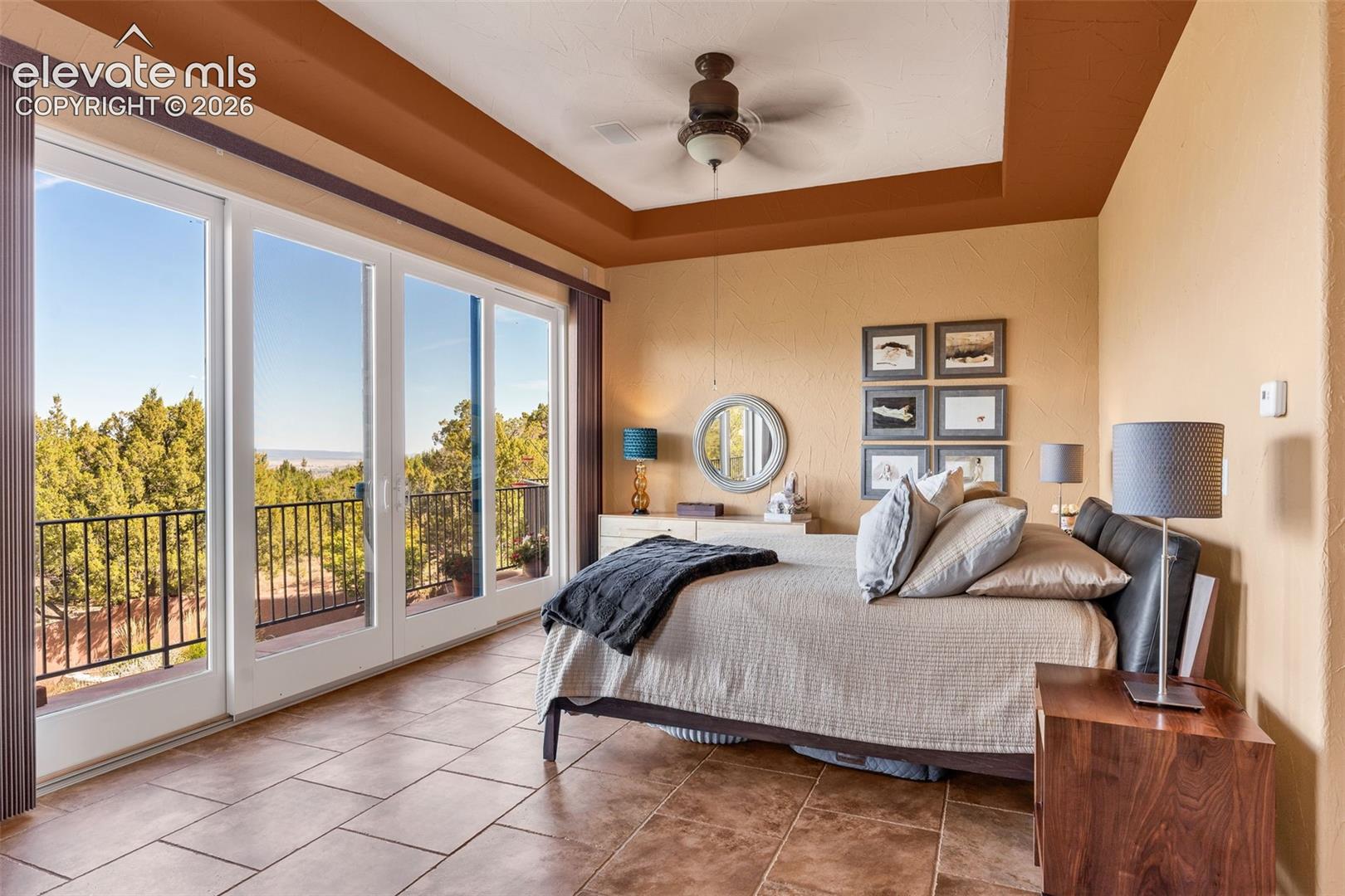 Trey ceiling with accent color, tile flooring, ceiling fan, and large sliding glass doors opening to an exterior balcony with metal railing
