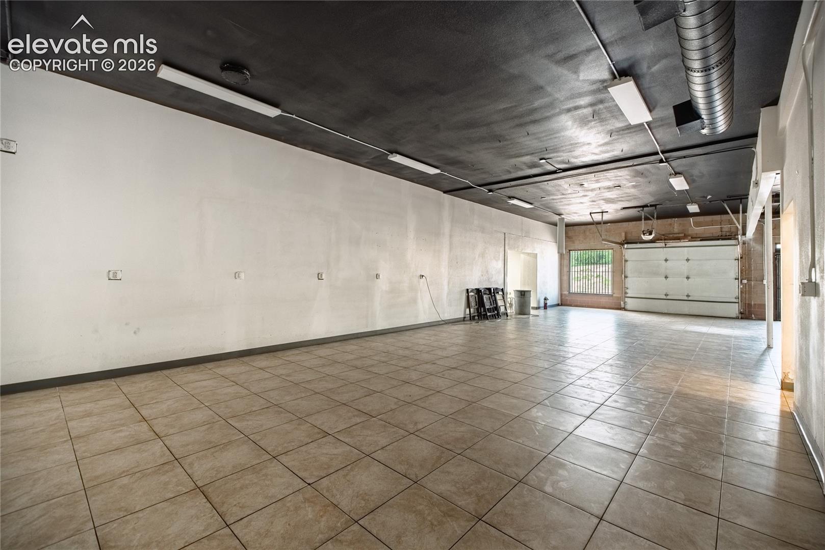 Spacious interior with tiled flooring, exposed ductwork, fluorescent lighting, and a large roll-up door