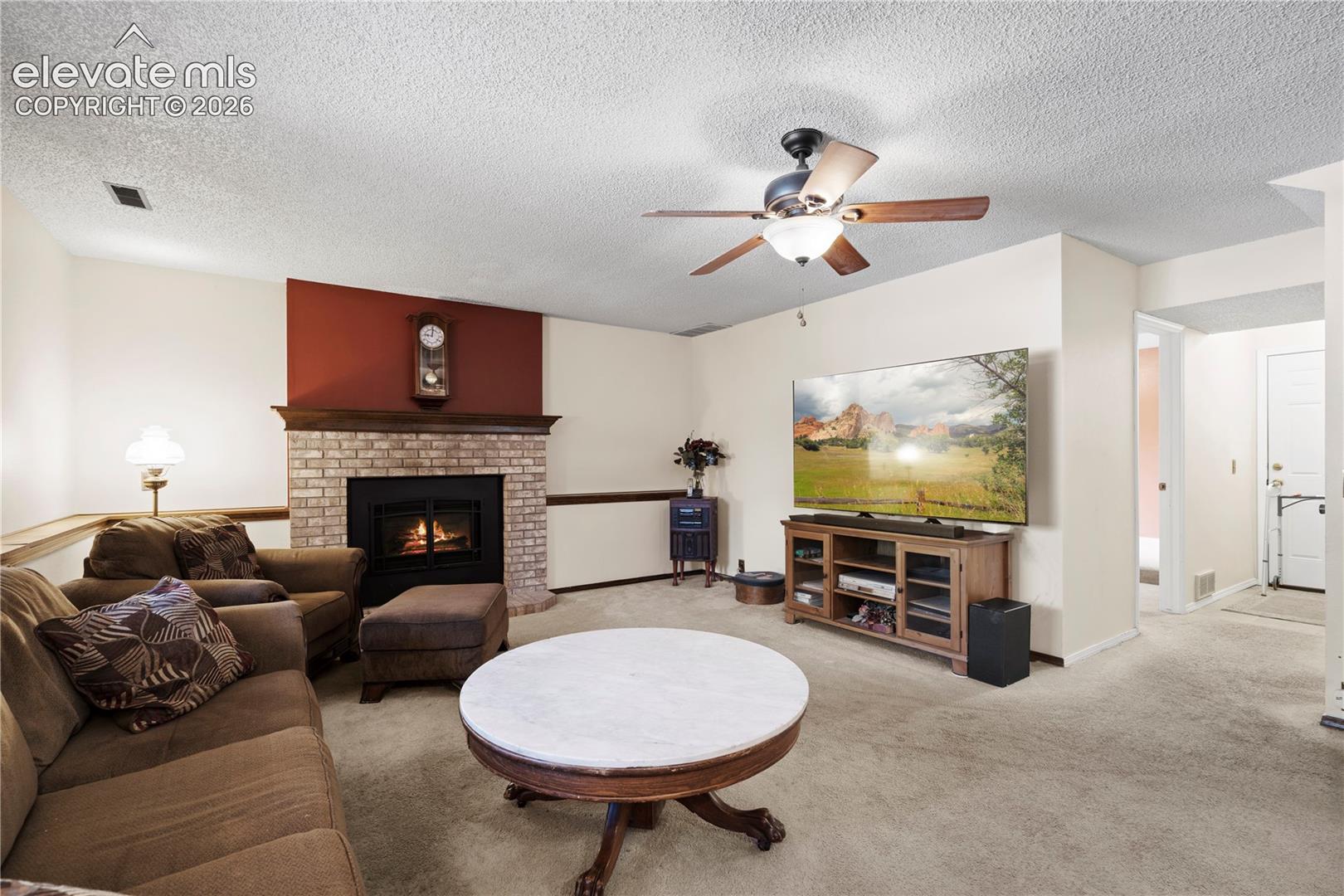 Living room featuring a brick fireplace with a wood mantle, ceiling fan with integrated lighting, and neutral carpeting
