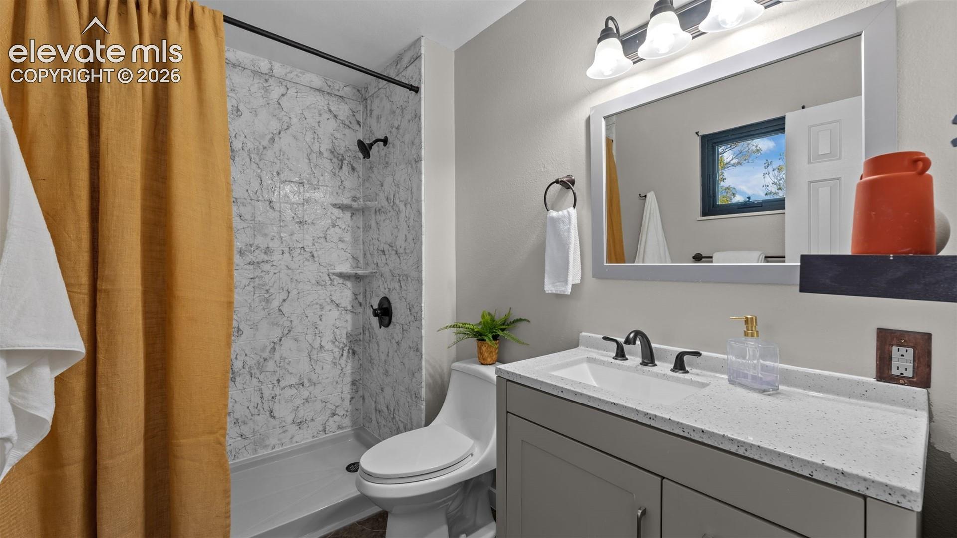Grey vanity cabinet with a speckled countertop, integrated sink, matte black faucet, and a tiled shower surround with built-in shelving