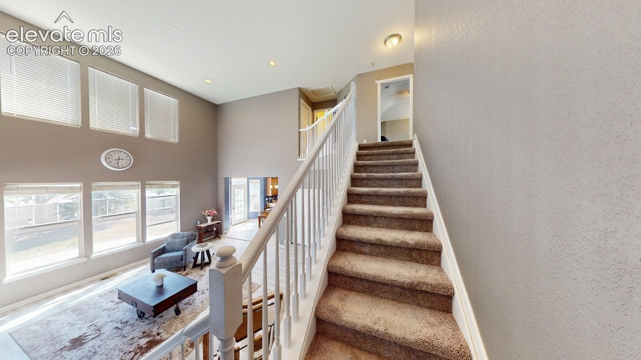 Two-story ceiling, multiple windows with horizontal blinds, overhead recessed lighting, and a white staircase with carpeted treads