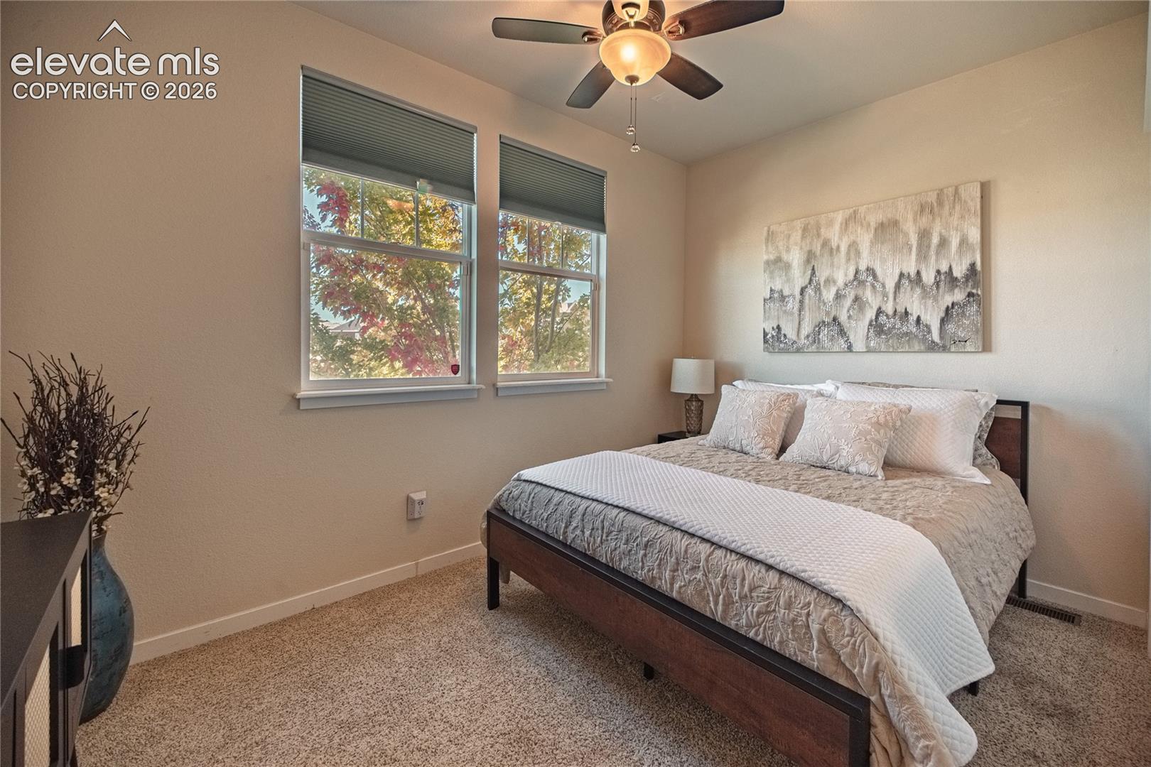 Carpeted bedroom featuring two large windows, a central ceiling fan with an integrated light fixture, and neutral wall tones