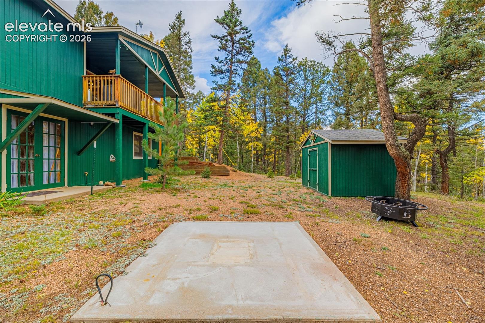 Exterior features a two-story wood-sided structure with an elevated wooden balcony, a detached storage shed, and a concrete pad, set on a lot with mature evergreen trees
