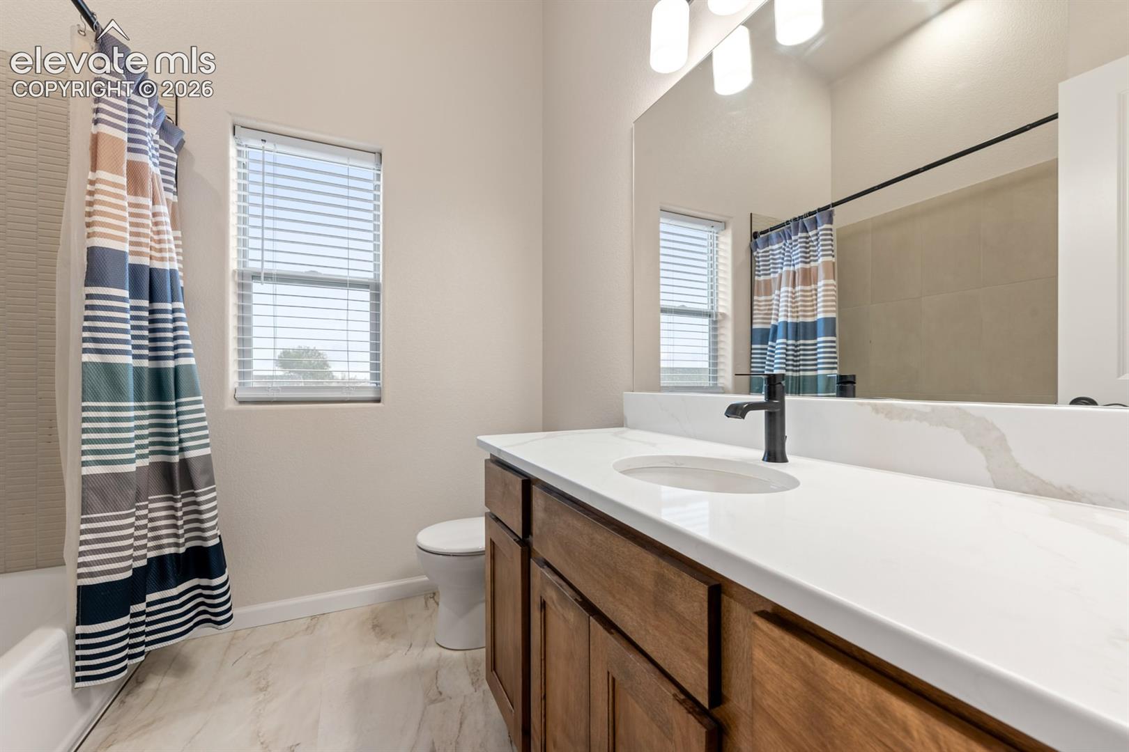 Bathroom featuring wood-finish cabinetry, white surface countertop with integrated sink, matte black faucet, full-width mirror, and stone-finish flooring