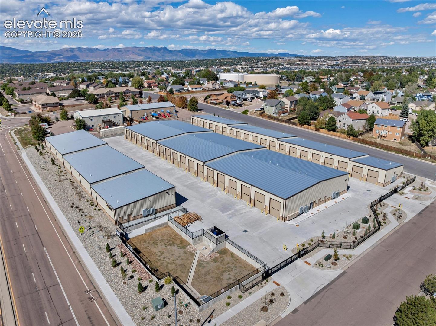 Multiple storage buildings with gray metal roofs, numerous roll-up doors, concrete paved access, and surrounding landscaping