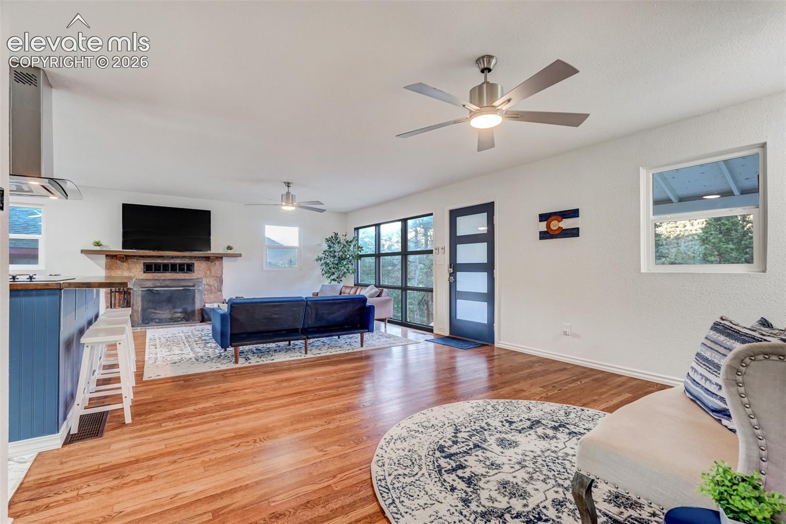Hardwood flooring, stone-clad fireplace, integrated range hood, modern ceiling fans, and a contemporary entry door with frosted glass panels