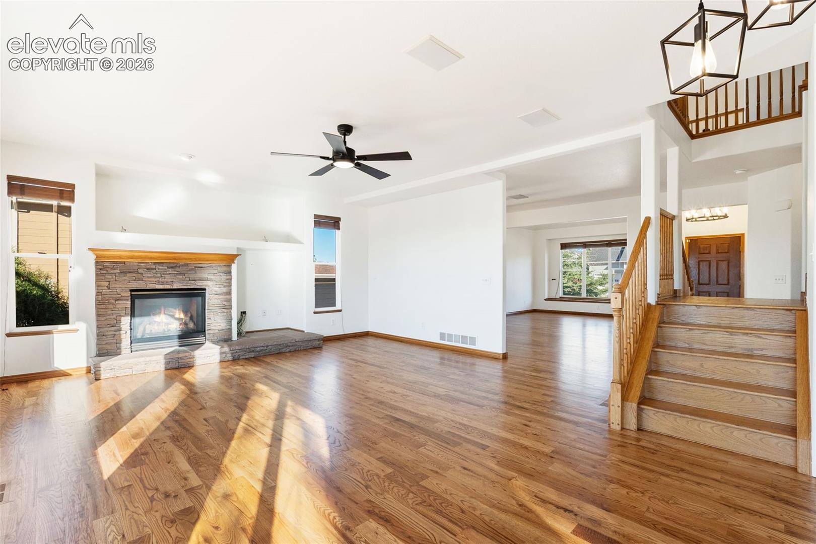 Living area featuring wood-finish flooring, stone-surround fireplace, wooden staircase with matching handrail, and geometric pendant lighting