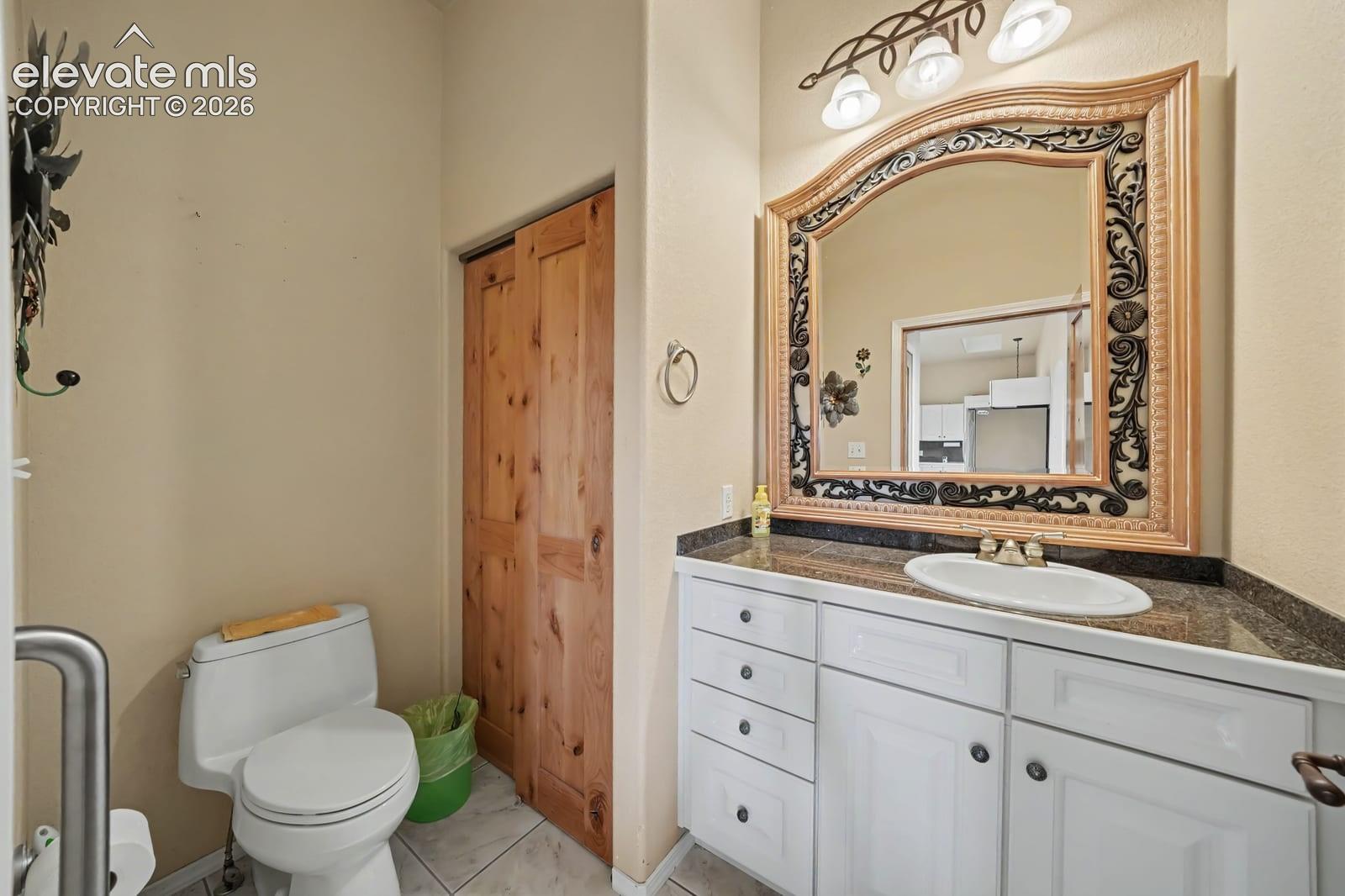Bathroom vanity featuring white cabinetry, stone-finish countertop with an oval drop-in sink, decorative framed wall mirror, and three-light overhead fixture, complemented by wood-finish bi-fold doors