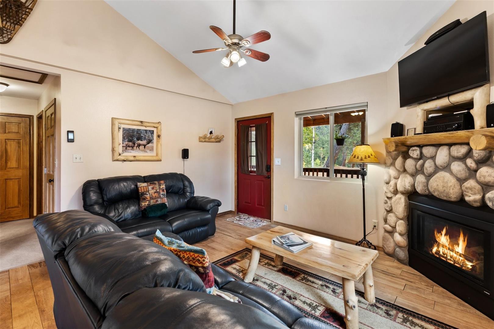 Vaulted ceiling with a ceiling fan, wood-finish flooring, stone-clad fireplace, built-in fireplace mantel, and a red entry door