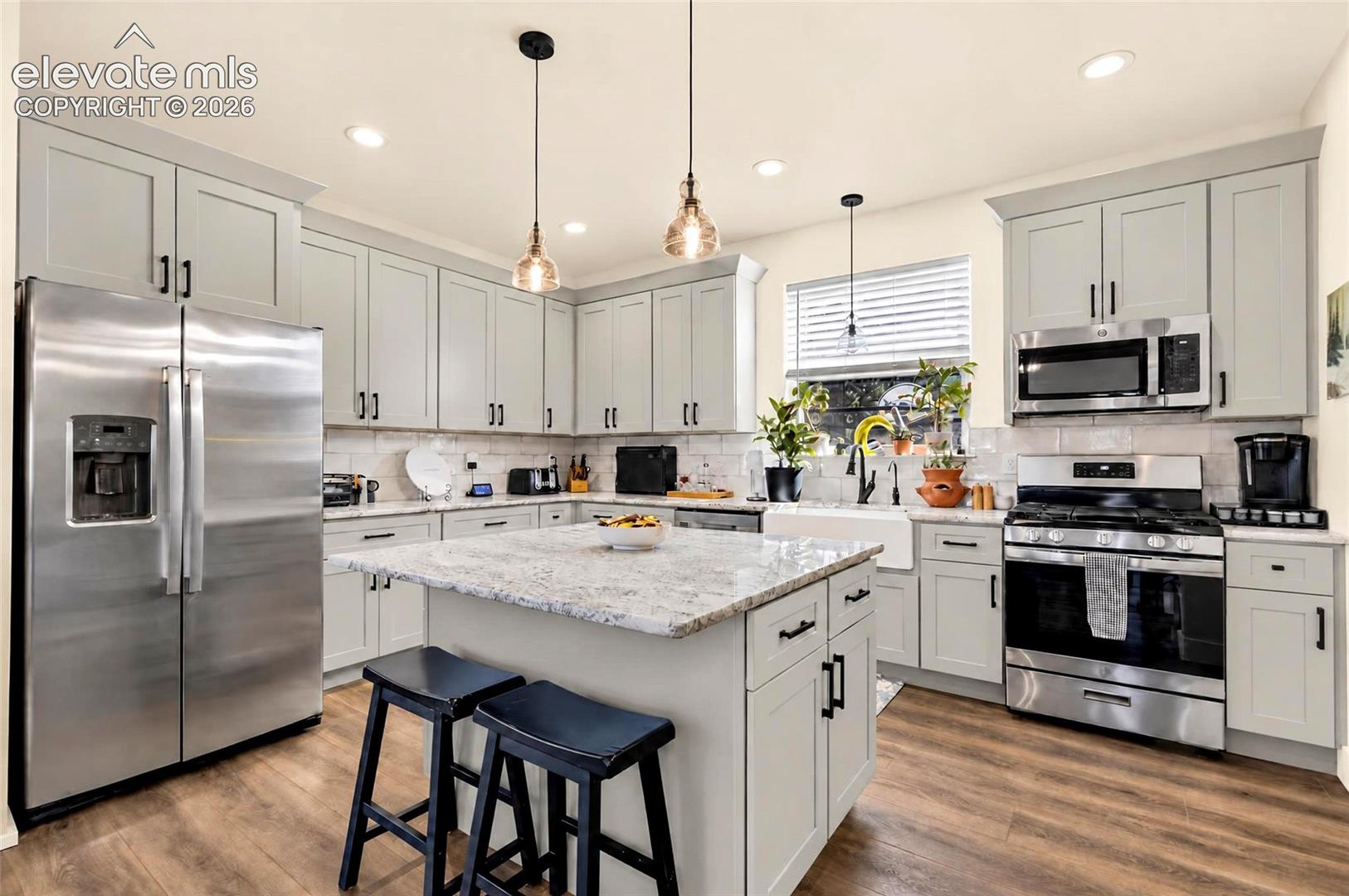 Kitchen featuring light grey Shaker-style cabinetry, a central island with stone-finish countertops, stainless steel appliances, a white farmhouse-style apron sink, and wood-finish flooring