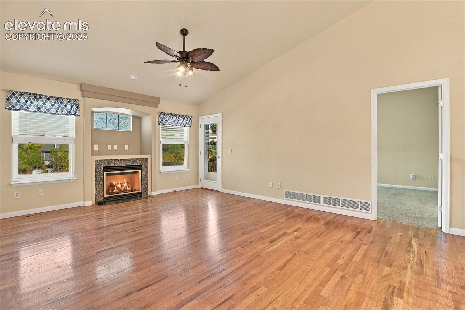 Living area featuring vaulted ceilings, wood-finish flooring, a stone-surround fireplace with built-in niche, and a multi-blade ceiling fan