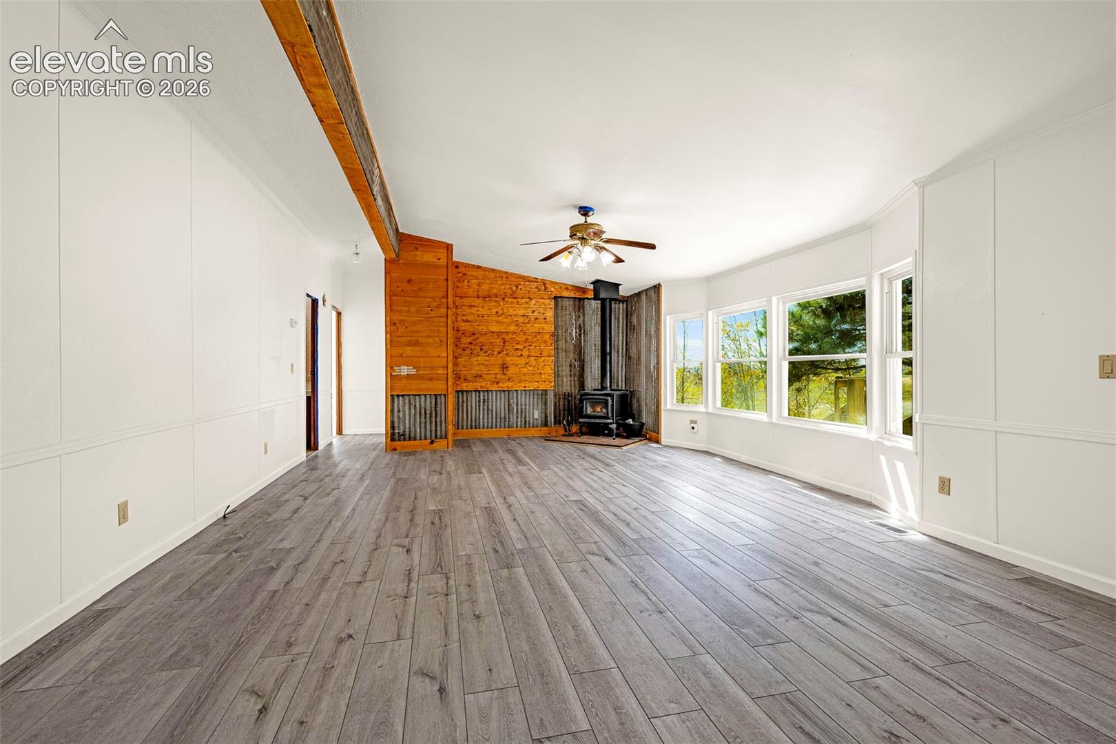 Living area featuring wood-finish flooring, vaulted ceilings with an exposed wooden beam, a wood-burning stove, corrugated metal accent wall, and expansive windows