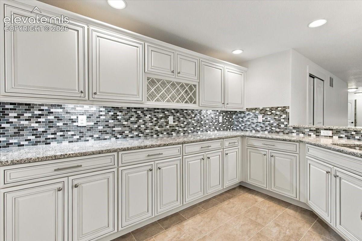 Kitchen featuring off-white cabinetry with shaker-style doors, stone-finish countertops, a multi-colored tile backsplash, integrated wine rack, and recessed ceiling lighting