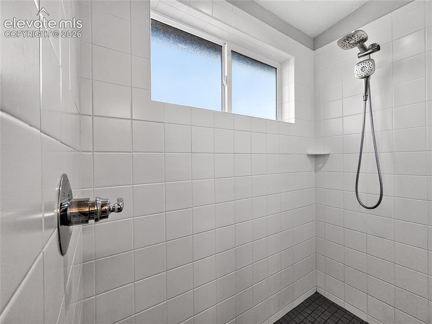 Shower featuring white tiled walls, a dual-head shower system, recessed shelving, and a textured dark floor in Master Bath