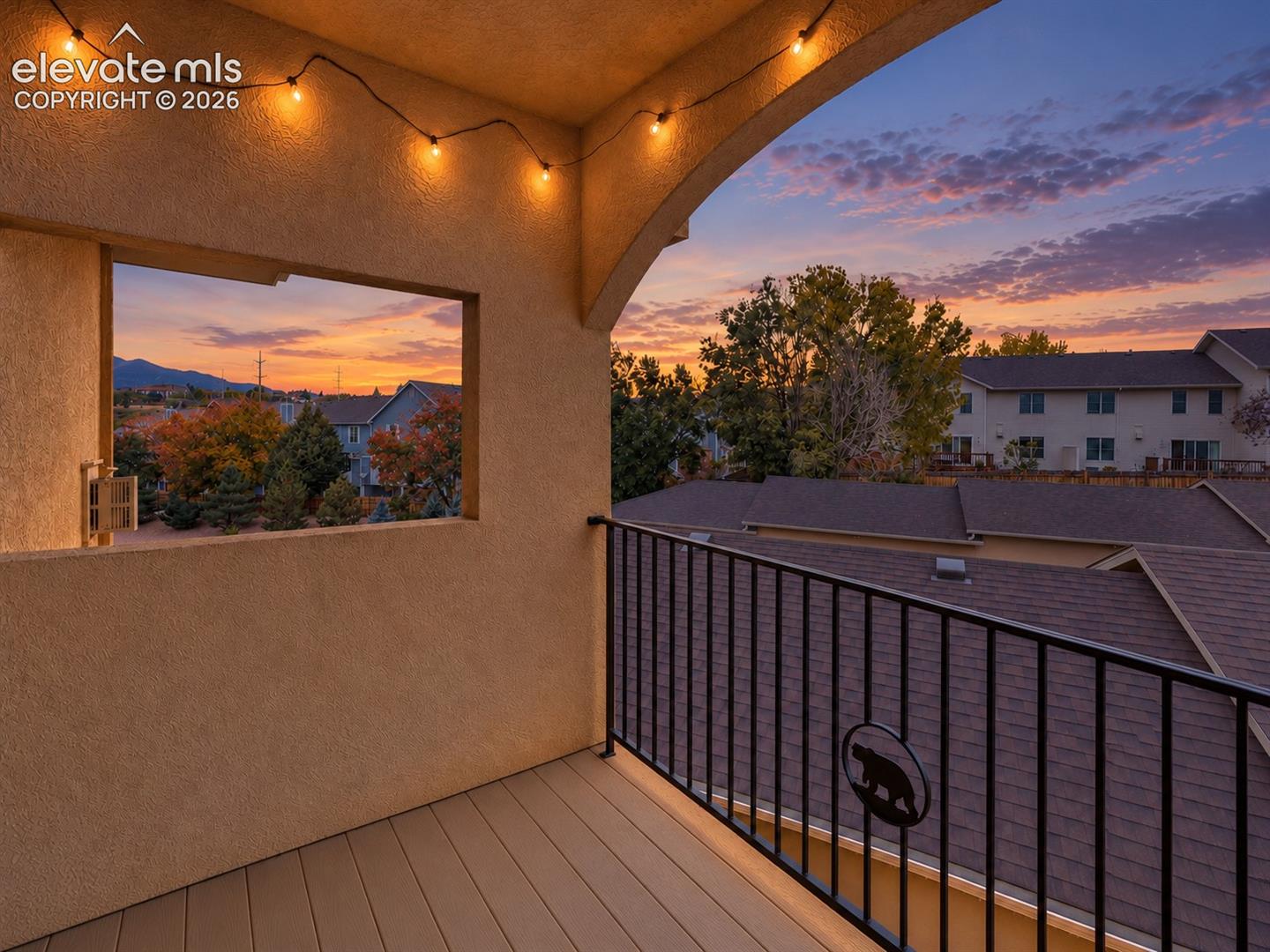 Covered balcony featuring stucco walls, composite decking, metal railing with decorative inlay, and integrated string lighting, complemented by a mountain-range backdrop