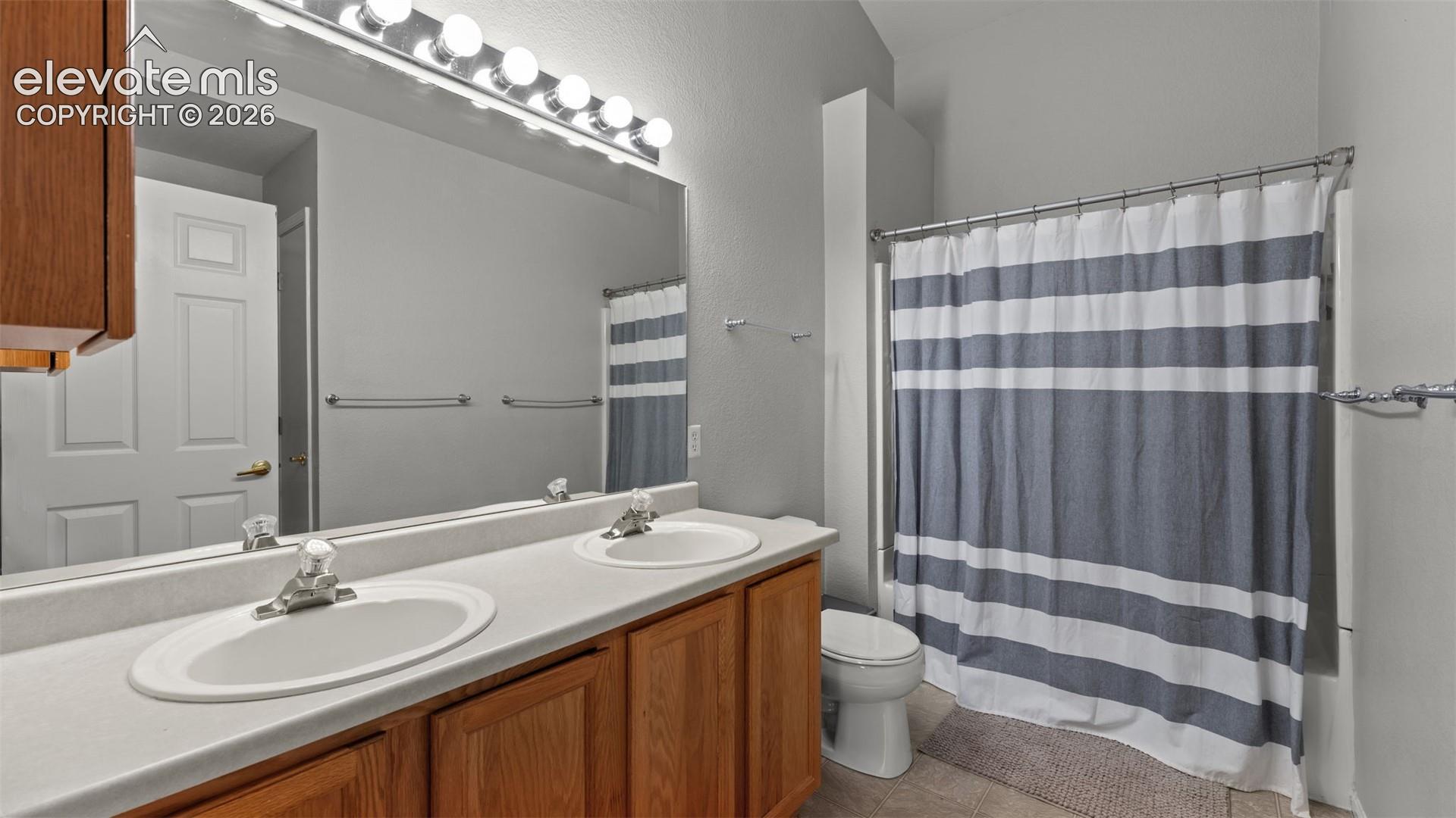 Bathroom featuring a dual-sink vanity with a solid-surface countertop, full-width vanity mirror, multi-bulb wall lighting, and a tub-shower combination