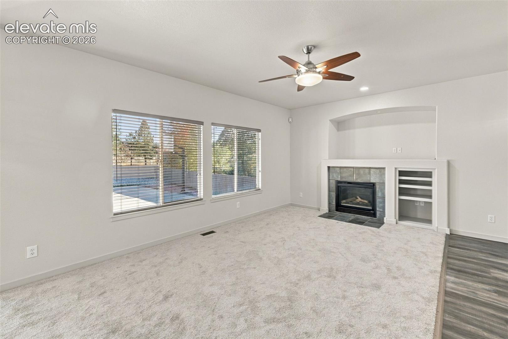 Living area featuring neutral carpeting, a fireplace with a stone-finish surround, built-in shelving, an arched recessed niche, and a multi-blade ceiling fan with integrated lighting