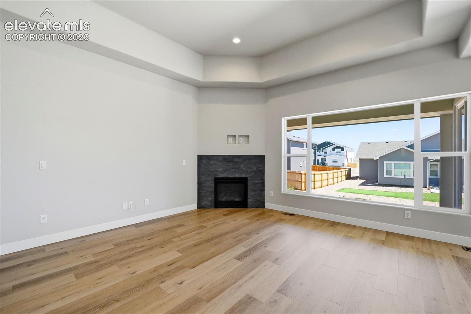 Living space featuring a tray ceiling, wood-finish flooring, corner fireplace with dark stone surround, and expansive windows