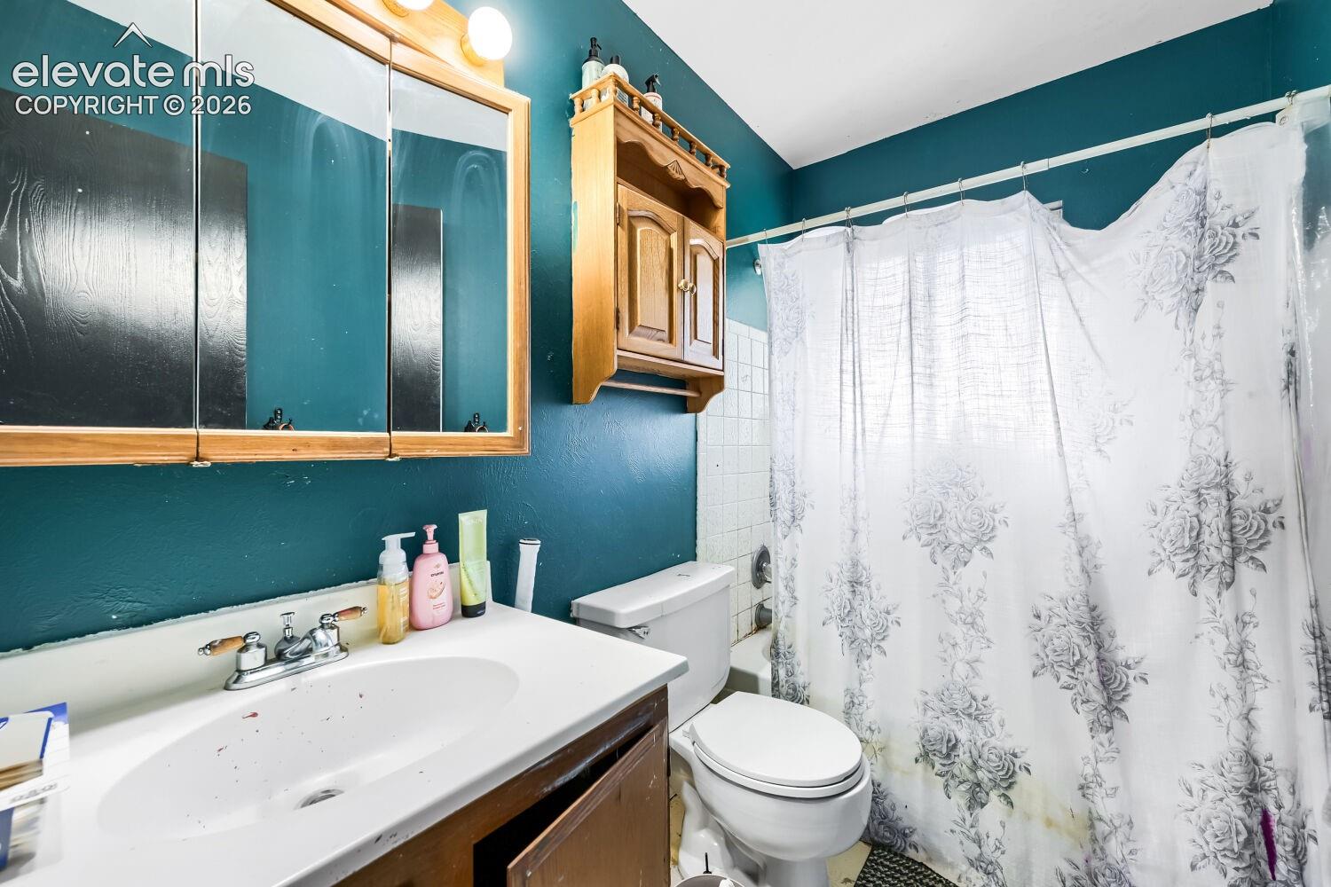 Bathroom featuring a vanity with an integrated sink, a medicine cabinet with mirrored doors, a wall-mounted wooden cabinet, a toilet, and a shower area with white tiled walls