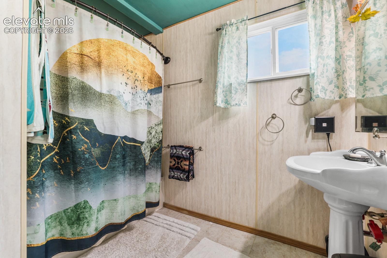 Bathroom featuring a pedestal sink with chrome faucet, a window with light-filtering curtains, and light-toned wall paneling