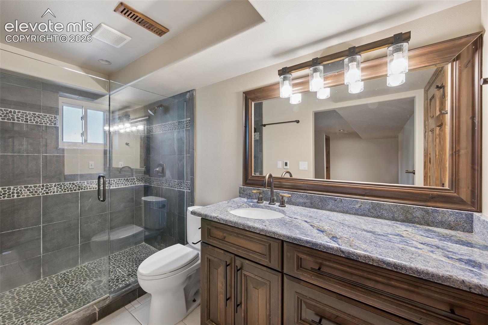 Basement bathroom featuring a dark wood-finish vanity with a stone countertop, an integrated sink, a glass-enclosed walk-in shower with stone tile detailing, and a large-format vanity mirror with integrated lighting
