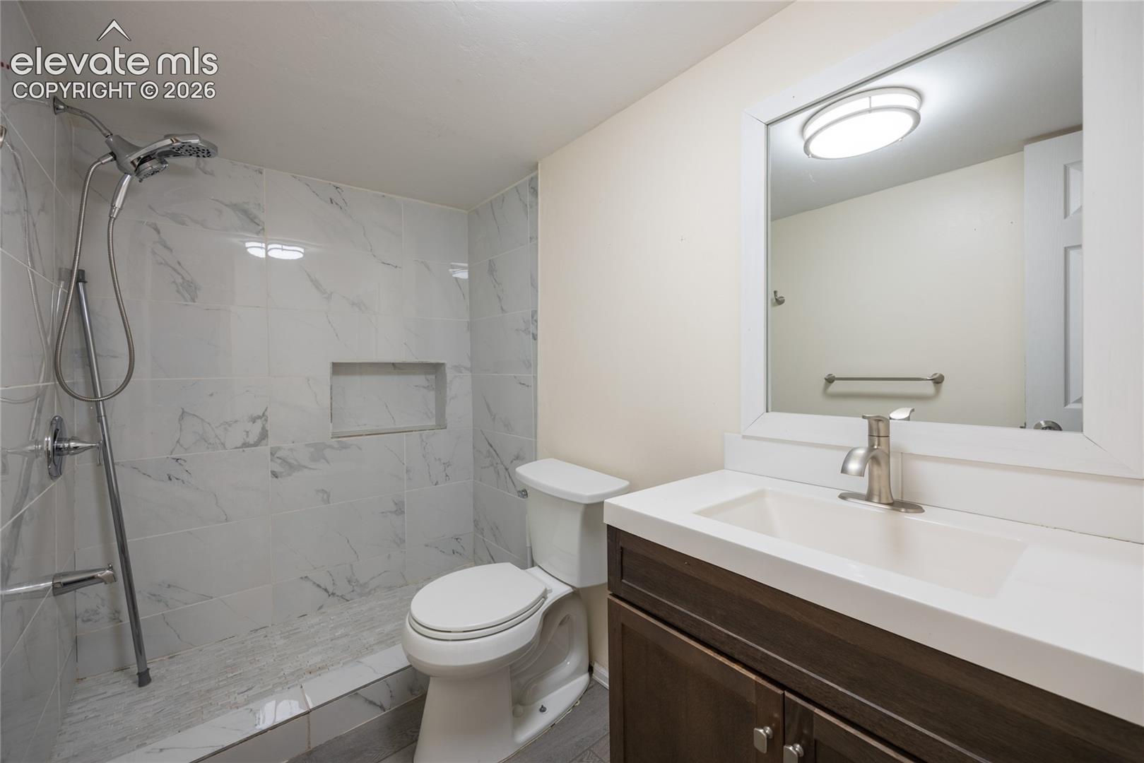 Walk-in shower with white marble-look tile, built-in shower niche, dark wood-finish vanity with white countertop, and rectangular undermount sink