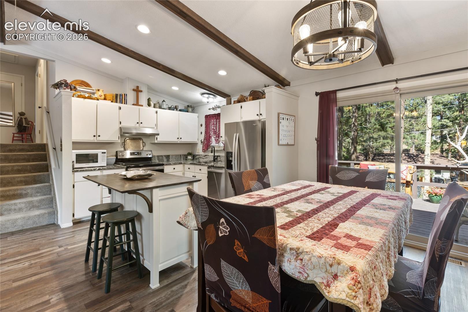 Open-concept kitchen and dining area featuring wood-finish flooring, exposed ceiling beams, white cabinetry, and stainless steel appliances