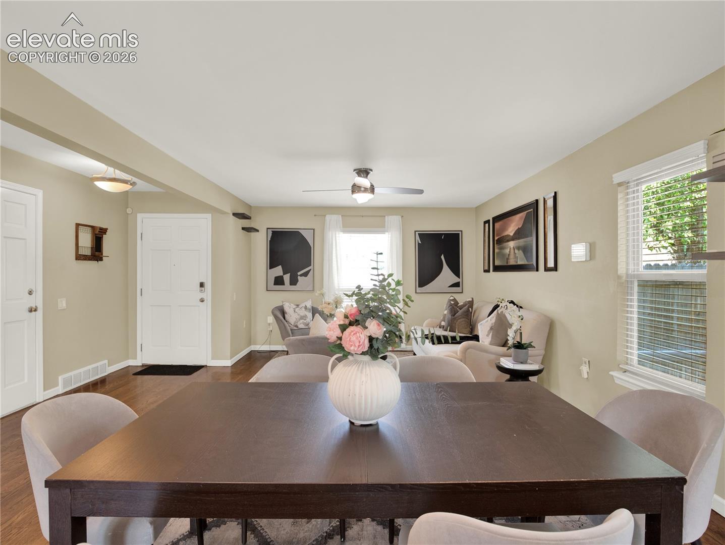 Open-concept living space featuring wood-finish flooring, a central ceiling fan with integrated lighting, and multiple windows with horizontal blinds