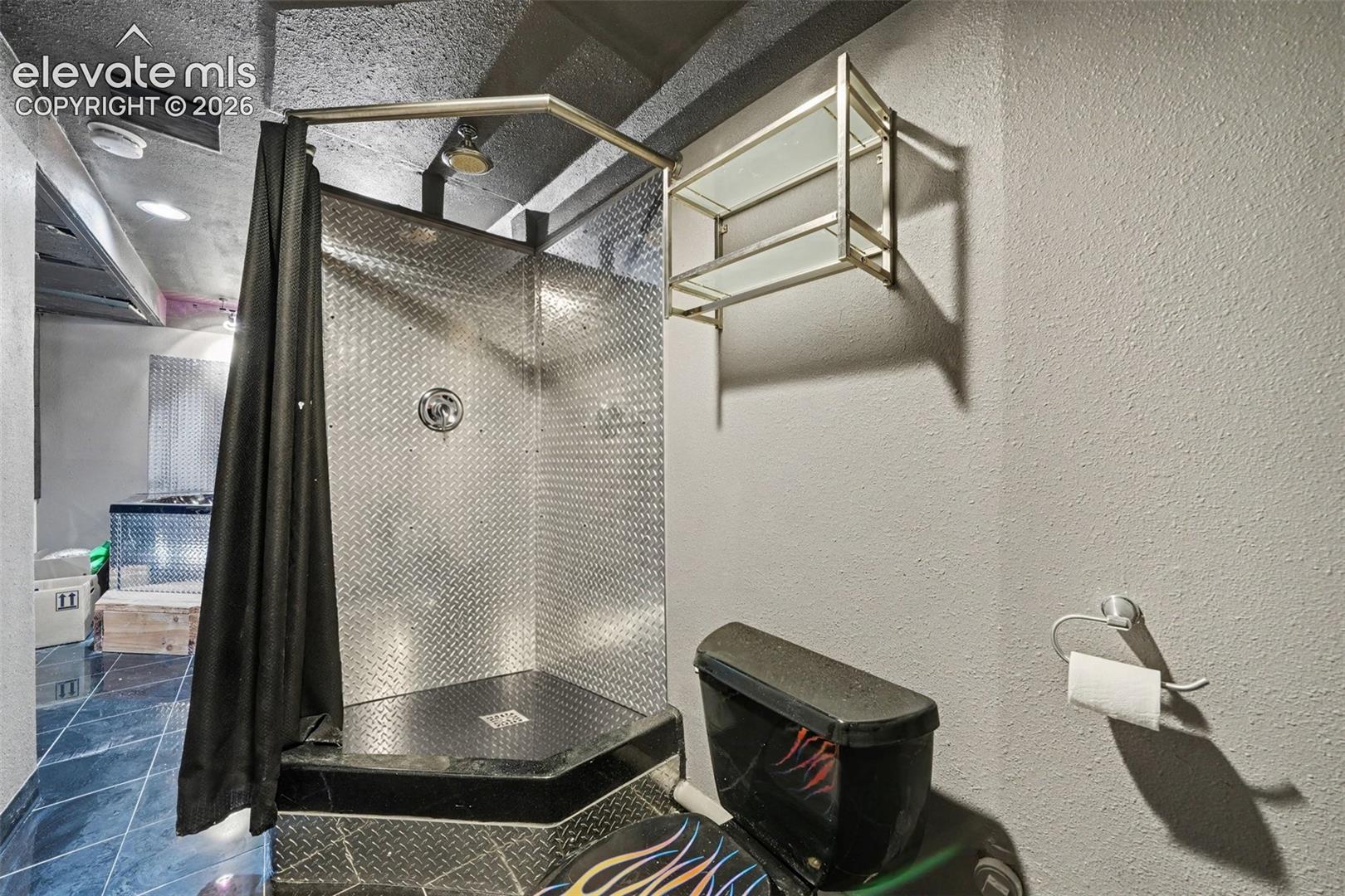 Shower enclosure featuring diamond plate metal wall paneling, a dark-toned tiered corner base, and a wall-mounted glass and metallic shelving unit