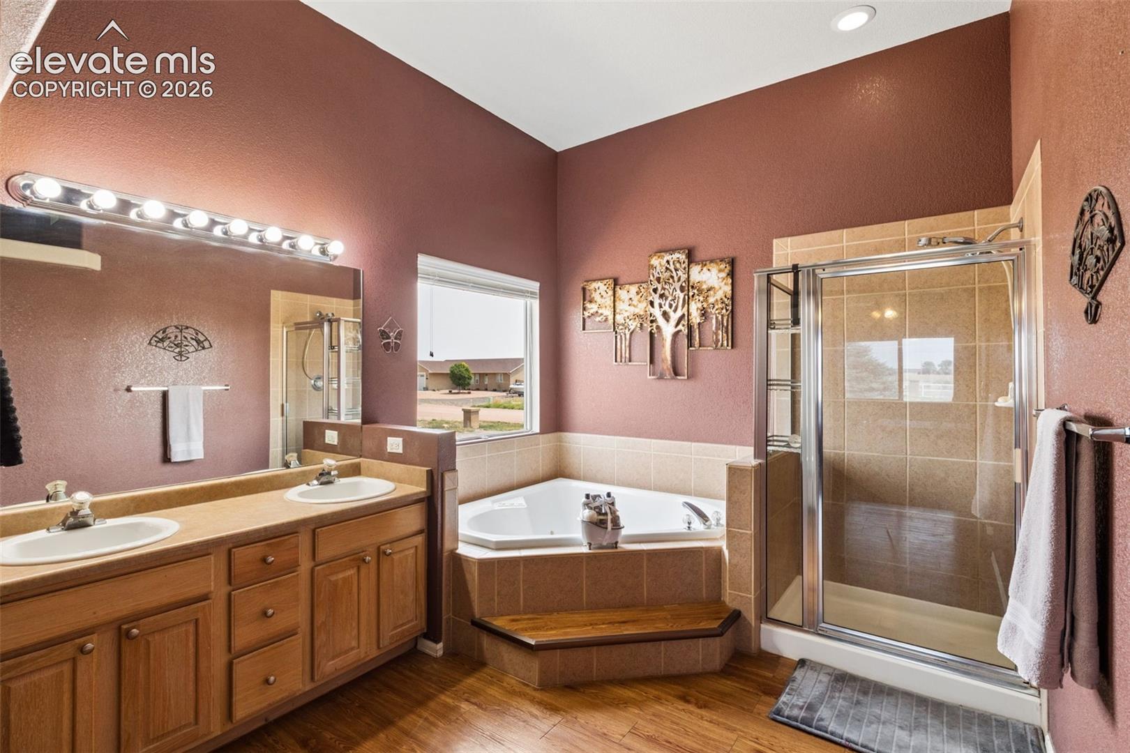 Bathroom featuring a dual-sink vanity with a large wall-mounted mirror, corner soaking tub with a wood-accented step, and a glass-enclosed shower with tiled surround, finished with wood-finish flooring and recessed lighting