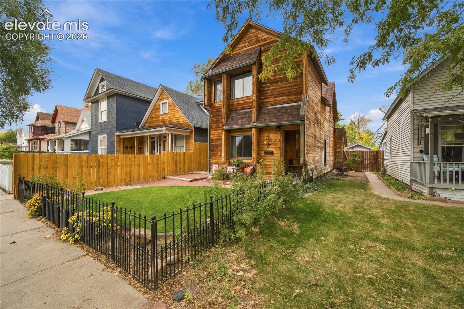 Two-story residence with wood siding, asphalt shingle roof, enclosed front yard with metal fencing, and brick-paved patio area