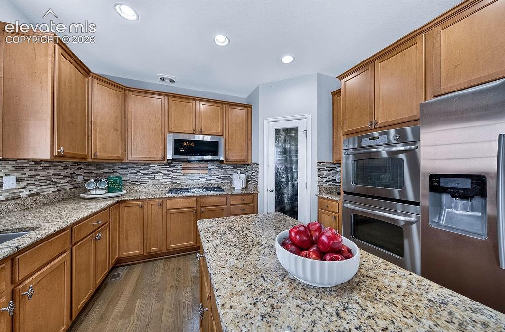 Kitchen featuring abundant wood-finish cabinetry, granite countertops, a mosaic tile backsplash, stainless steel appliances, and wood-finish flooring