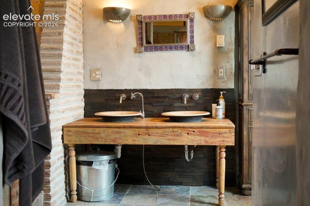 Dual vessel sinks set on a rustic wood-finish vanity, complemented by a mosaic-framed mirror and stone-finish wall sconces