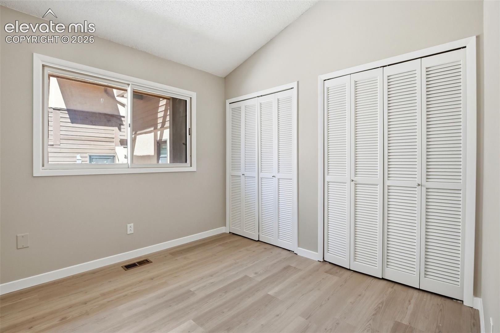 Interior room featuring wood-finish flooring, two sets of louvered bifold closet doors, a horizontal sliding window with white trim, and a vaulted ceiling