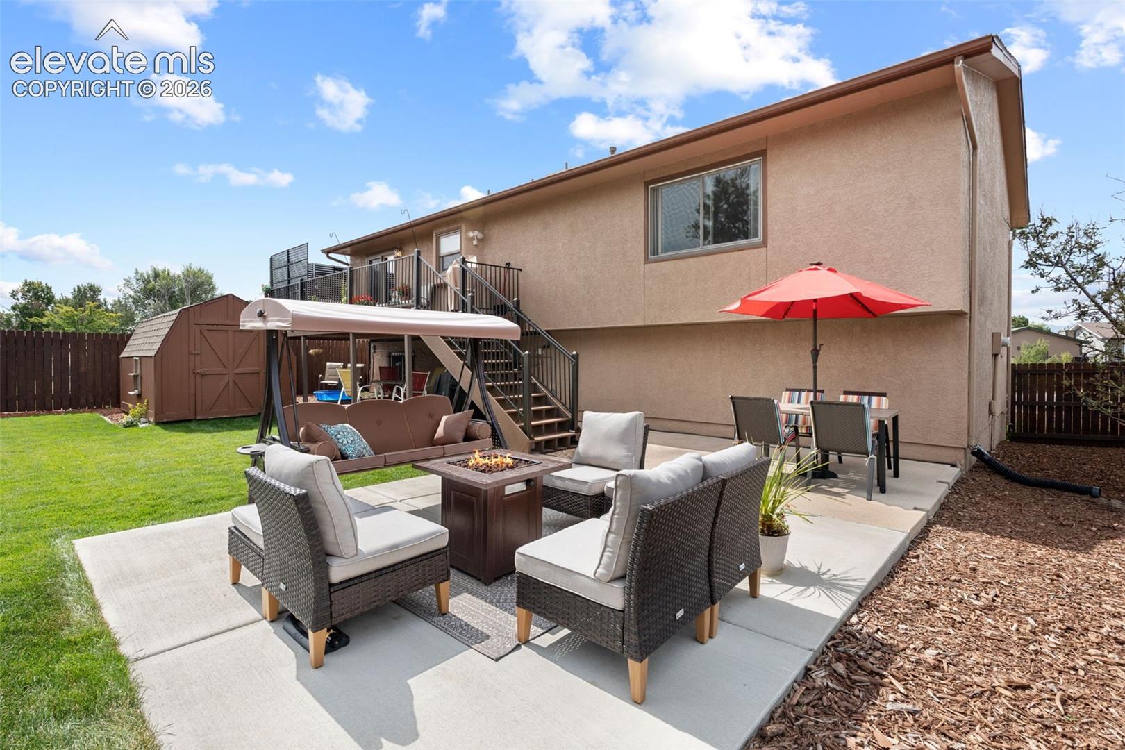 Stucco-exterior residence featuring an expansive concrete patio, elevated staircase with metal railing, and detached storage shed within a fenced yard