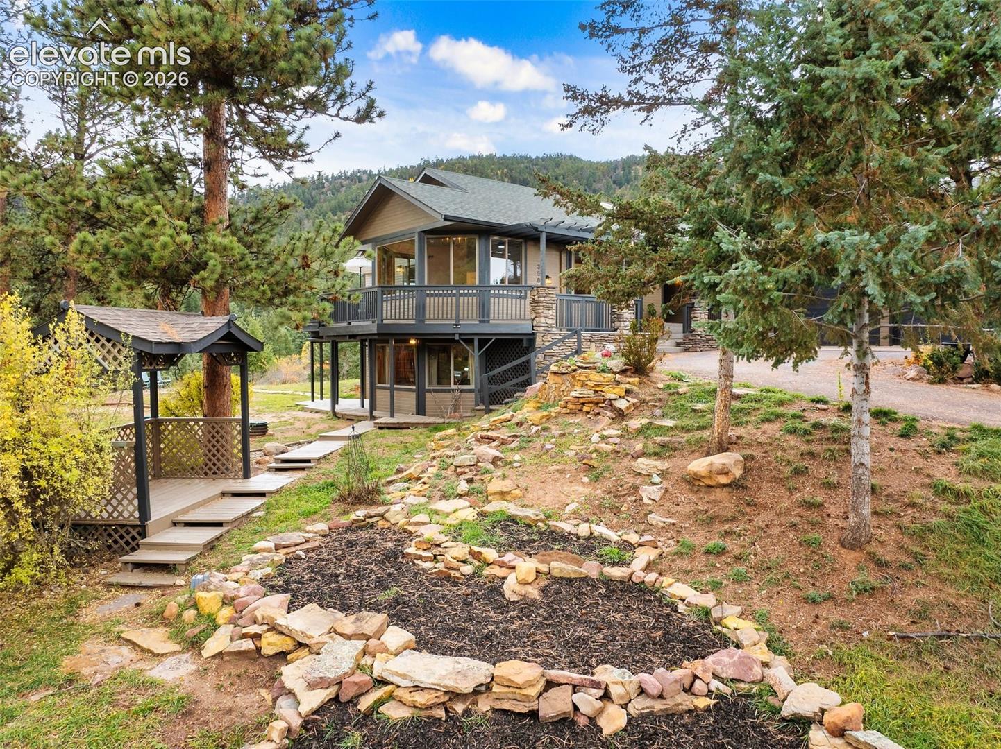 Custom-built residence featuring stone masonry accents, an elevated wraparound deck with railing, a detached covered pergola, natural stone terracing, and mature evergreen landscaping