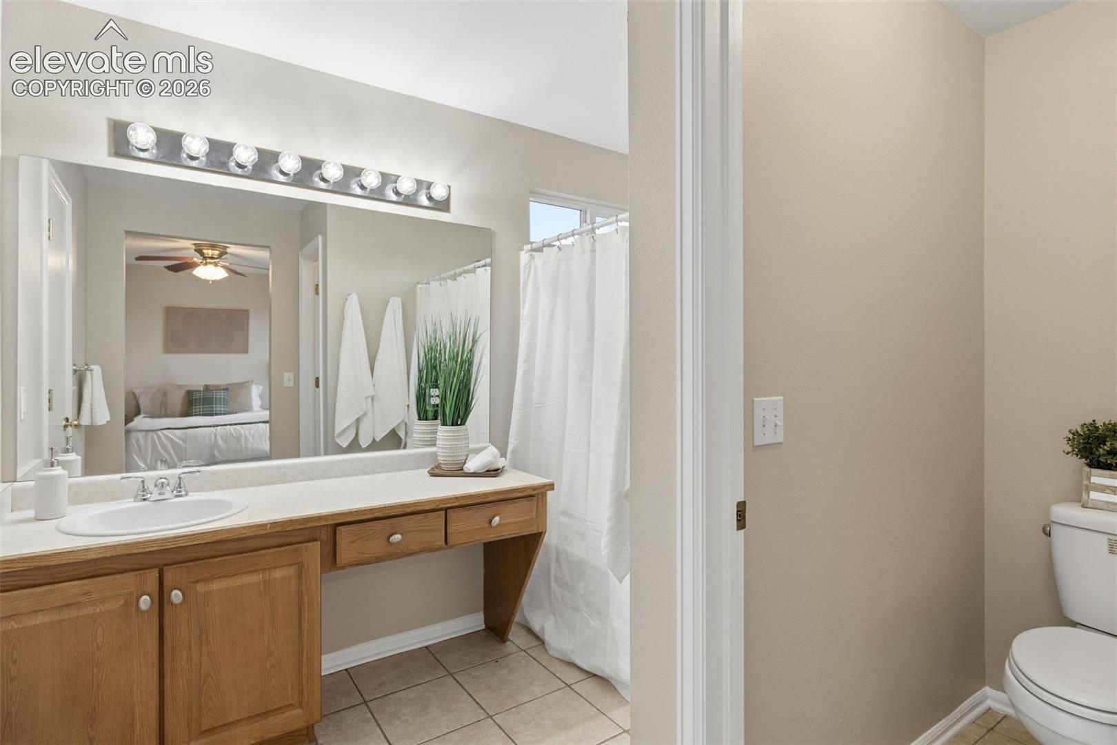 Bathroom featuring a vanity with wood-finish cabinetry, integrated countertop with a rounded basin, oversized wall-mounted mirror, multi-bulb overhead vanity lighting, and ceramic tile flooring
